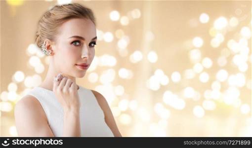jewelry, luxury and people concept - portrait of woman in white dress with diamond earring and finger ring over beige background and festive lights. woman in white dress with diamond jewelry