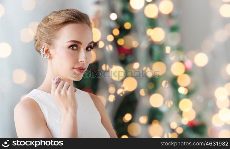jewelry, luxury, holidays and people concept - smiling woman in white dress with diamond earring and ring over christmas tree lights background