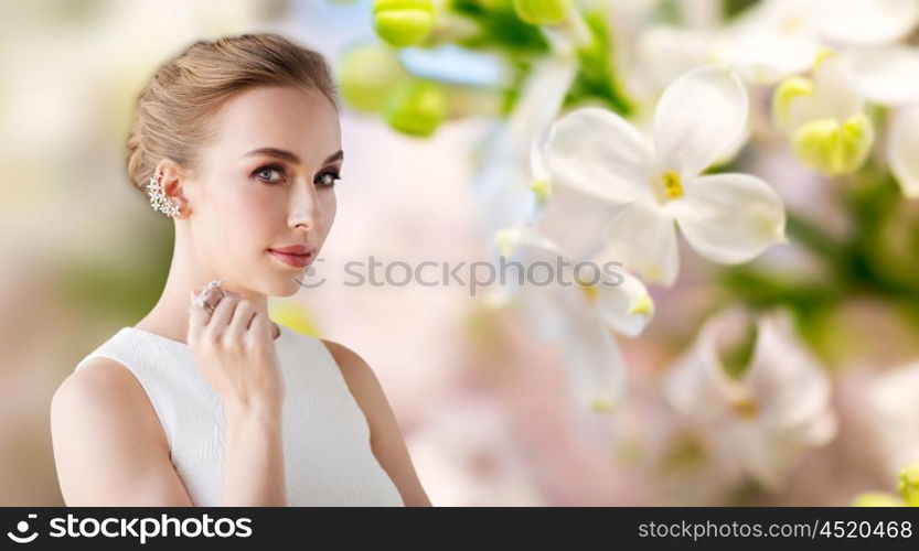 jewelry, luxury, wedding and people concept - smiling woman in white dress with diamond earring and ring over natural lilac blossom background