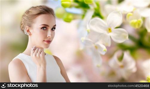 jewelry, luxury, wedding and people concept - smiling woman in white dress with diamond earring and ring over natural lilac blossom background