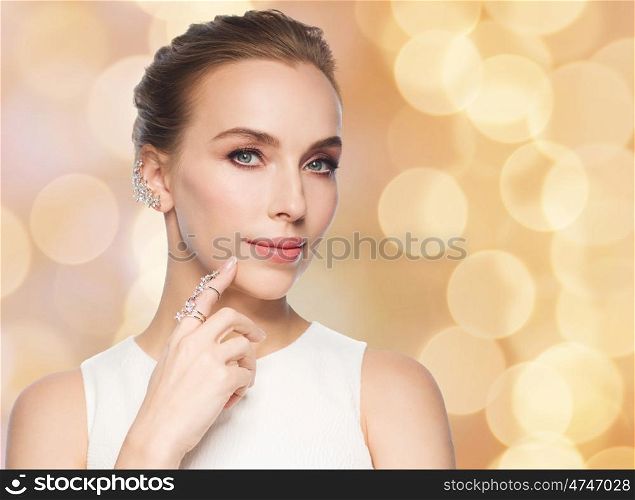 jewelry, luxury, wedding and people concept - smiling woman in white dress with diamond earring and ring over beige holidays lights background