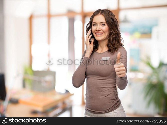 job, work and business concept - happy woman with name tag calling on smartphone and showing thumbs up over office background. happy woman calling on smartphone at office