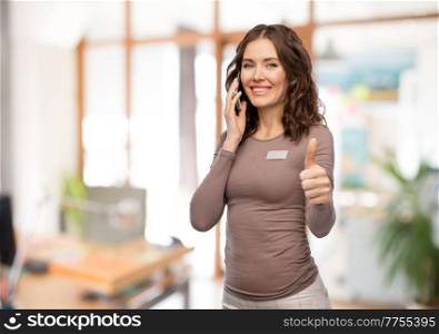 job, work and business concept - happy woman with name tag calling on smartphone and showing thumbs up over office background. happy woman calling on smartphone at office