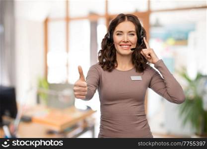 job, work and customer service concept - happy female helpline operator or woman with headset and name tag showing thumbs up over office background. woman with headset showing thumbs up at office