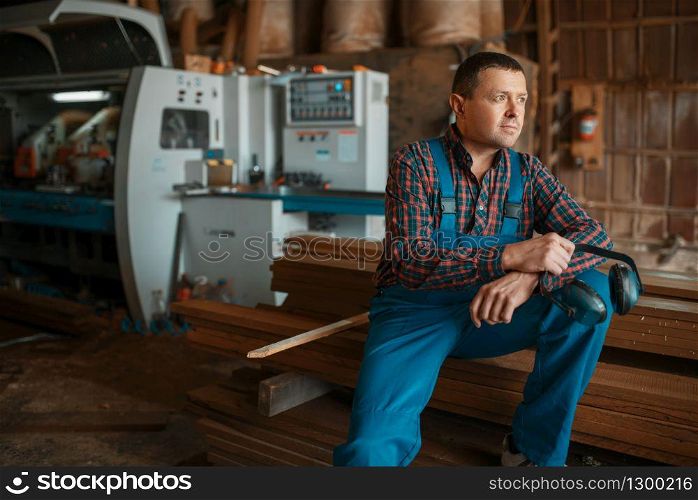 Joiner in uniform at his workplace on timber mill, woodworking machine on background, lumber industry, carpentry. Wood factory, forest sawing in lumberyard. Joiner in uniform at his workplace on timber mill