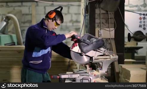 Joiner wearing protective goggles and earmuffs cutting wooden plank with sliding mitre saw in workshop. Carpenter using manual circular saw for wood in workplace.