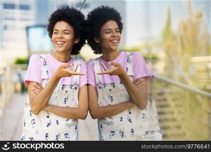 Joyful African American female gesturing and looking away with smile while standing near glass wall with reflection on street against blurred background. Laughing black woman near glass building