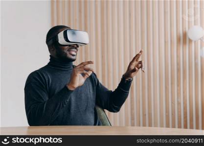 Joyful African man in VR headset glasses playing favorite 3D game while sits at desk at home office smiling and waving with hands up and down touching air. Innovative technology concept. Young joyful African man office worker in VR headset glasses playing favorite 3D game