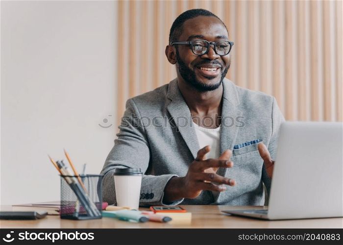 Joyful Afro american businessman wearing glasses and stylish blazer making video call, looking at screen with bright smile, positive entrepreneur leading remote meeting, participating online conference. Joyful Afro american businessman wearing glasses and stylish blazer making video call on laptop