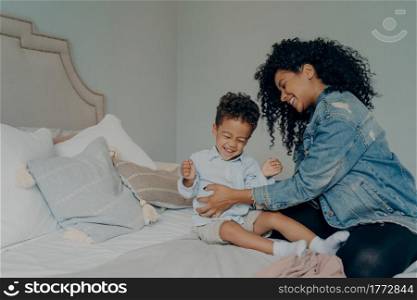 Joyful afro american mother with bright smile and curly hair tickling her cute little son toddler while spending happy time with each other in bedroom, loving mom and child having fun, playing indoor. Loving afro american mother tickling son while having fun together in bedroom