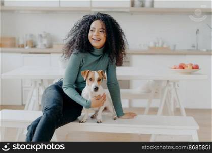 Joyful Afro woman sits at white bench together with dog against kitchen interior, table with plate full of red apples, get pleasure while playing at home. Animal owner feels care and responsibility