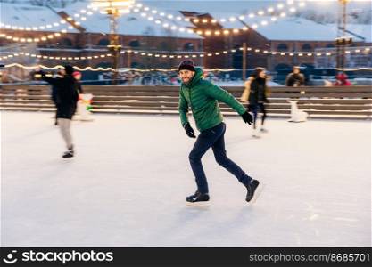 Joyful bearded man practices going skating on ice ring, has cheerful expression, smiles happily, demontrates his professionalism. Active sporty male in green jacket go in for winter sport