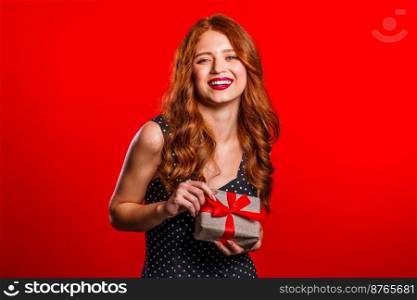 Joyful beautiful woman with perfect makeup holding gift box with bow on red wall background. Retro styled girl smiling, she is glad to get present. Joyful beautiful woman with perfect makeup holding gift box with bow on red wall background. Retro styled girl smiling, she is glad to get present.