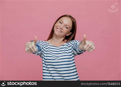 Joyful beautiful young lady raises thumbs at camera, shows that everything is okay, wears striped clothes, tilts head, smiles broadly, shows white teeth, isolated over pink background, says yes