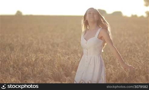 Joyful blond female in white dress spinning around in golden wheat field in glow of beautiful sunset in summer. Excited woman with flying long hair enjoying freedom and happiness while spending leisure in countryside at sunset. Slow motion.