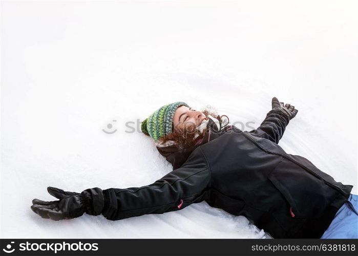 Joyful cheerful girl lying down on the white clean snow with raised up hands and making snow angel, enjoying snowy weather, happy winter holidays