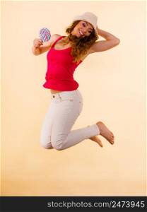 Joyful crazy woman wearing red tshirt white trousers jumping while holding lollipop candy in hand. Happiness fun concept. Studio shot on bright beige. Positive woman jumping holds lollipop candy in hand