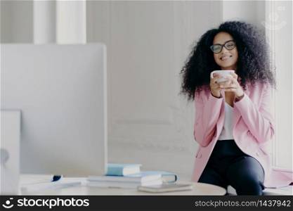 Joyful curly haired female freelancer or blogger has coffee break, holds white mug with beverage, poses near window, focused in computer, watches useful training video, wears near formal clothes