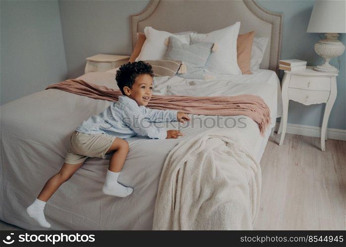 Joyful cute little mixed race kid with bright smile trying to climb on big beige bed with lots of pillows while playing alone in bedroom at home, sweet toddler boy enjoying playtime indoors. Joyous little african american kid enjoying playtime indoors on bed