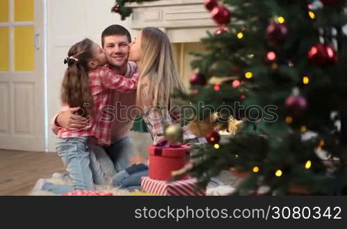 Joyful daughter and attractive mother kissing their handsome father over Christmas decorated interior. Portrait of affectionate family with little cute girl spending great time together on xmas eve at home. Dolly shot.