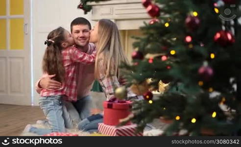 Joyful daughter and attractive mother kissing their handsome father over Christmas decorated interior. Portrait of affectionate family with little cute girl spending great time together on xmas eve at home. Dolly shot.