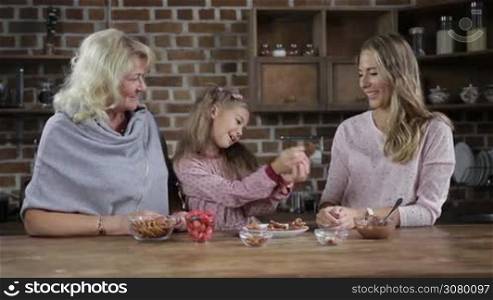 Joyful daughter giving homemade chocolate cookies to beautiful mother and smiling grandmother in the kitchen. Cheerful little girl sharing delicious cookies with her loving mom and grandma while multi generation family spending leisure at home.