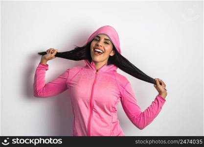 Joyful ethnic female model dressed in sports jacket touching hair and shouting happily in studio against white background while looking at camera. Happy Hispanic woman in bright sportswear in studio