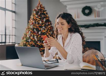 Joyful europian woman sitting at desk with laptop, reading xmas news or message on smartphone while working remotely in room with christmas decoration, happy female shopping online via mobile phone. Joyful europian woman sitting at desk with laptop, reading xmas news or message on smartphone