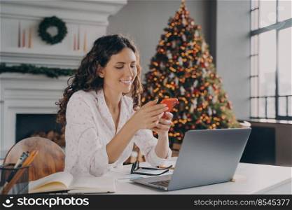 Joyful europian woman sitting at desk with laptop, reading xmas news or message on smartphone while working remotely in room with christmas decoration, happy female shopping online via mobile phone. Joyful europian woman sitting at desk with laptop, reading xmas news or message on smartphone