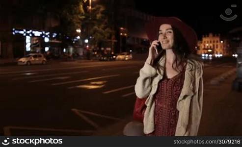 Joyful fashionable brunette female in bordo hat talking on smartphone as she walks on the sidewalk in night city. Smiling young woman communicating on cellphone while walking along city street in the evening. Steadicam stabilized shot