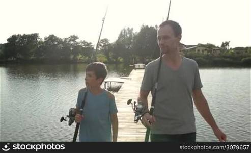 Joyful father and excited son with fishing rods walking on wooden pier for fishing on lake in glow of sunset. Carefree dad and teenage boy going fishing by calm pond on sunny summer day as they spend leisure together. Slo mo. Stabilized shot.