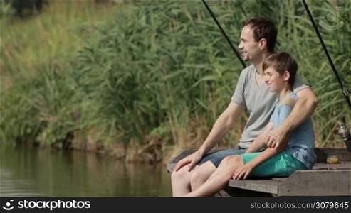 Joyful father embracing his teenage son with love and tenderness while sitting on wooden pier near the pond and fishing in summer. Caring dad and boy bonding with each other while spending weekend by the lake, fishing together. Slow motion.