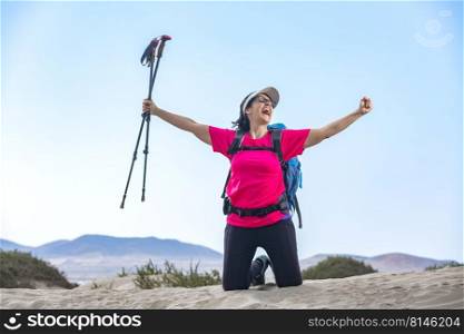 Joyful female backpacker with trekking poles celebrating success with outstretched arms on Famara beach and looking away. Delighted traveler celebrating victory on seashore