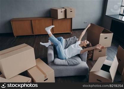 Joyful female homeowner having fun in armchair lifting hands celebrates relocation to new home in room with cardboard boxes. Happy woman tenant feels success on moving day. Mortgage, first realty.. Joyful woman having fun in armchair celebrates relocation to new home. Mortgage, first realty