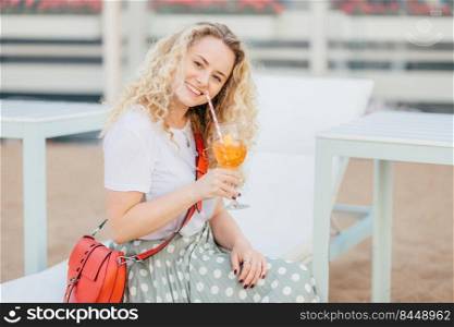 Joyful female model with friendly smile, healthy pure skin, holds glass of cocktail, wears stylish clothes, recreats on beach during summer vacation, being in good mood. People and lifestyle concept