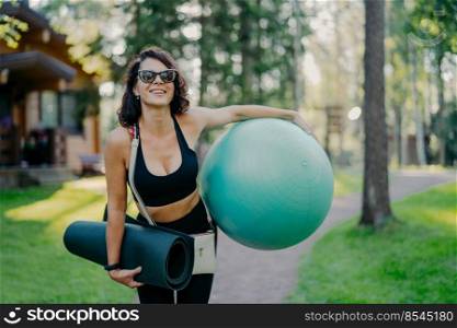 Joyful female smiles broadly, holds big fitness ball, rolled up karemat, wears sunglasses, enjoys summer day, goes in for sport regularly, being pilates instructor, poses outdoor at beautiful nature