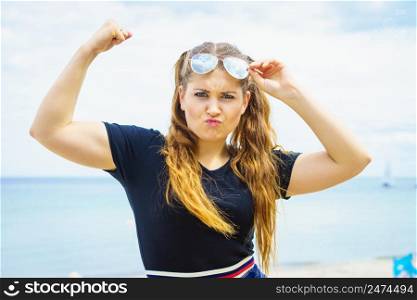 Joyful funny woman showing off her arm muscles biceps. Female wearing sunglasses during summertime.. Woman in sunglasses showing muscles