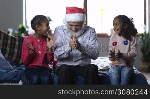 Joyful grandfather in santa hat with beard giving christmas gift boxes to his excited mixed race granddaughters at home. Two happy little sisters embracing their affectionate granddad on the bed as family celebrating christmas in domestic room.