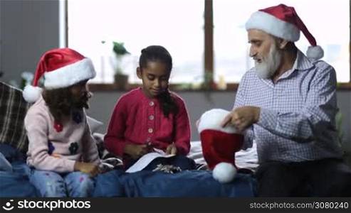 Joyful grandpa with beard in santa hat and lovely mixed race granddaughters get ready to play secret santa on christmas. Cute girl with scissors cutting papers with names, her sister wrapping them and putting into santa hat held by granddad.