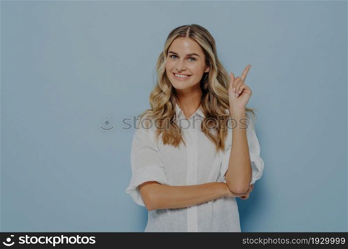 Joyful happy young blonde female with wavy long hair pointing with forefinger up with expression just look at this, isolated at blue wall with copy space for text or advertising. Promotion concept. Joyful happy young blonde female with wavy long hair pointing with forefinger up
