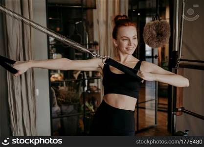 Joyful happy young fit woman pilates instructor doing hand strengh exercise on cadillac reformer, stretching arms and smiling during pilates training in gym. Concept of healthy lifestyle. Joyful young woman pilates instructor doing hand strengh exercise on reformer