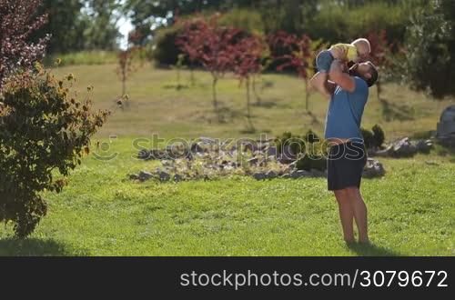 Joyful hipster father in casual clothes throwing his infant boy high up in the air over beautiful landscape background. Happy dad and toddler baby son lifting him up, playing and having fun in summer park while enjoying leisure together in sunlight.