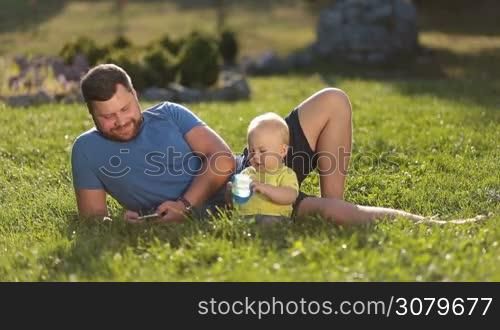 Joyful hipster father with beard surfing the net on smartphone while spending leisure in nature together with adorable infant son. Handsome dad and little toddler baby boy relaxing on green park lawn on summer sunny day.