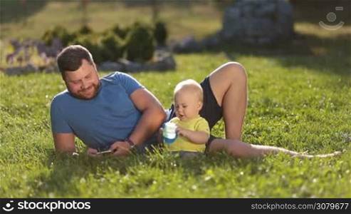 Joyful hipster father with beard surfing the net on smartphone while spending leisure in nature together with adorable infant son. Handsome dad and little toddler baby boy relaxing on green park lawn on summer sunny day.