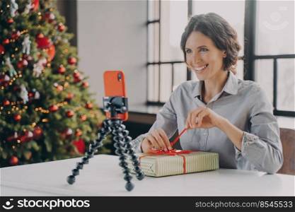 Joyful lady sitting at table with Xmas tree on background in front of phone on tripod, smiling blogger streaming and sharing tips for decorating christmas gifts or home interior with followers online. Joyful lady sitting at table with Xmas tree on background in front of phone on tripod