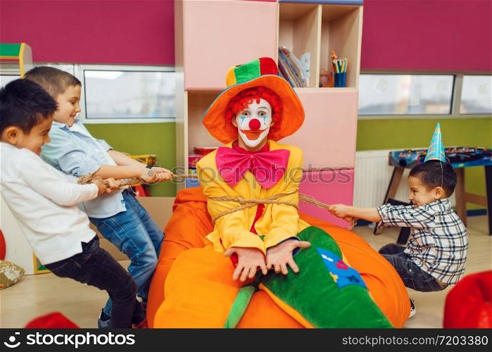 Joyful little boys tied up funny clown in children&rsquo;s area. Birthday party celebrating in playroom, baby holiday in playground. Childhood happiness, childish leisure. Joyful little boys tied up funny clown
