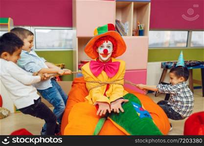 Joyful little boys tied up funny clown in children&rsquo;s area. Birthday party celebrating in playroom, baby holiday in playground. Childhood happiness, childish leisure. Joyful little boys tied up funny clown