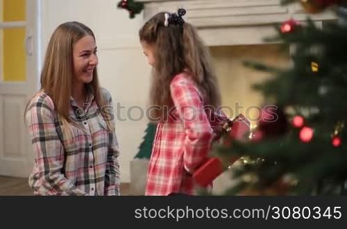 Joyful little girl surprising her attractive mother with Christmas present and kissing her. Adorable daughter giving xmas gift to her charming mother while family enjoying winter holidays in festive decorated room. Dolly shot.