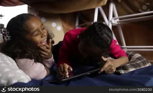 Joyful little mixed race sisters watching funny video on digital tablet while lying down in cubby house made of blanket and chairs. Excited cute girls having fun and playing with touchpad at home. Dolly shot. Slow motion.