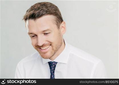 Joyful male corporate worker in elegant white shirt, looks happily down, remembers something pleasant in mind, has toothy smile, wears elegant white shirt with tie, isolated over white wall.